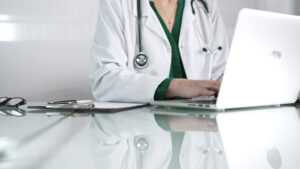 Doctor woman using technology in healthcare, close up of hands typing on laptop with stethoscope, glasses, and clipboard on glass desk. Medicine concept