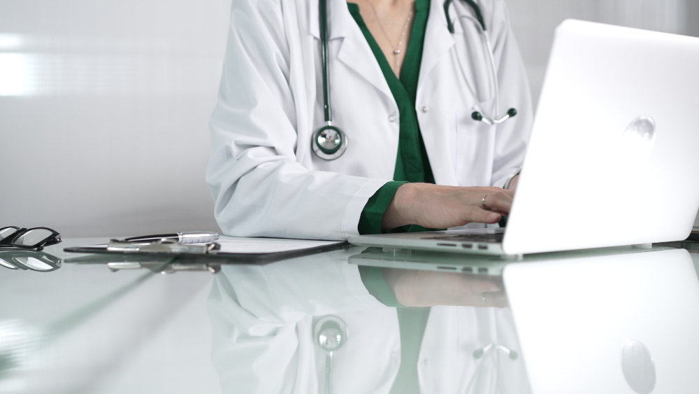 Doctor woman using technology in healthcare, close up of hands typing on laptop with stethoscope, glasses, and clipboard on glass desk. Medicine concept